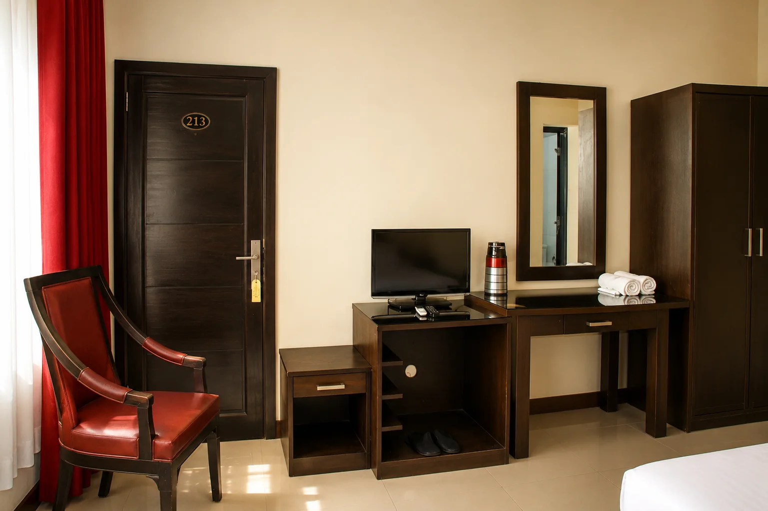 Fasilitas interior Superior Room dengan pintu kamar, kursi, TV, meja, cermin, lemari, handuk, dan lantai keramik.